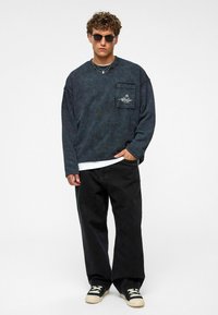 Vervaagd donkerblauw sweatshirt met een voorzak en wit logo; gecombineerd met losse zwarte broek en zwart- en crèmekleurige sneakers. Geen zichtbare patronen.