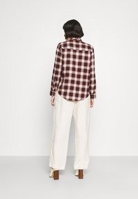 Karierter Shirt in Rot und Weiß mit abgerundetem Saum, langen Ärmeln und geknöpften Manschetten, kombiniert mit locker sitzenden weißen Leinenhosen und cremefarbenen Absatzschuhen.