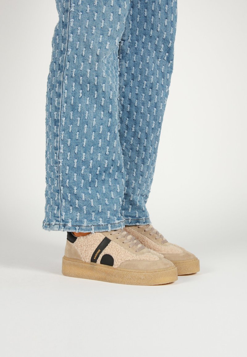 Beige sneakers with a textured finish, black accents, and tan soles, worn with light blue patterned denim jeans.
