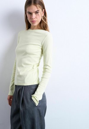 Woman wearing a light yellow long-sleeve top and loose dark gray pants, standing with one hand in pocket against a plain wall.