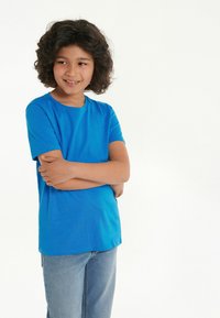 Dítě s kudrnatými tmavými vlasy v modrém tričku a džínách stojí s crossed arms, usmívá se a dívá se do strany na pozadí bez vzoru.