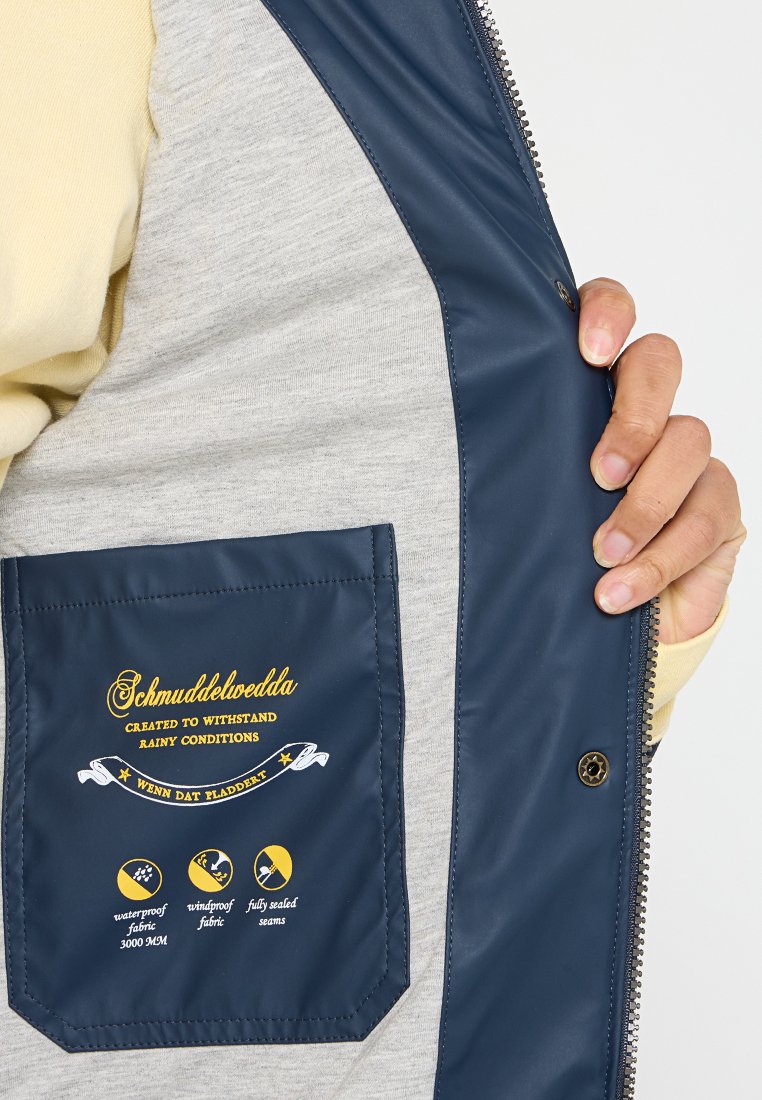 Gilet blu navy e grigio in materiale impermeabile, con un'etichetta stampata in giallo sulla tasca che evidenzia le proprietà impermeabili e antivento.