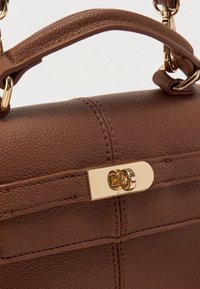 Καφέ δερμάτινη τσάντα χειρός με δομημένο σχέδιο, χρυσό εξοπλισμό και λεπτομερείς ραφές. Το κούμπωμα είναι μια ορθογώνια πλάκα με διπλές τρύπες.