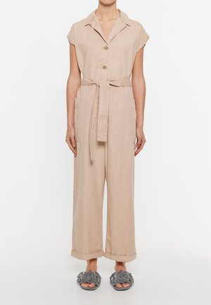 Beżowy lniany kombinezon z krótkimi rękawami, guzikami z przodu i paskiem w talii. Rozszerzane nogawki z bocznymi kieszeniami; zestawiony z szarymi kwiatowymi butami wsuwanymi.