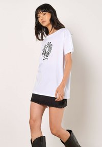 Weißes Baumwoll-T-Shirt mit einem schwarzen geometrischen Sonnenmotiv, lässiger Schnitt und kurzen Ärmeln, kombiniert mit einem schwarzen Jeansrock und schwarzen Stiefeln.