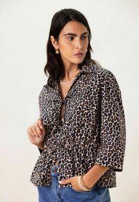 Blouse à imprimé léopard avec un col à boutons, une taille peplum et des manches trois-quarts, fabriquée en tissu doux. Accentuée par des liens subtils.