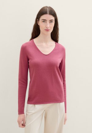 Pull à manches longues en V en tissu rose doux, avec une texture lisse et une coupe décontractée, associé à un pantalon crème.