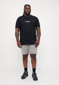 Zwarte t-shirt met korte mouwen met witte tekst "Original Studio", gecombineerd met grijze shorts en zwarte sneakers. Model staat rechtop tegen een effen achtergrond.