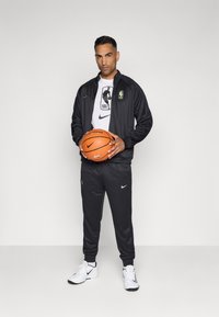 Črn Nike trenirni komplet z zelenimi detajli, bela NBA logo majica, drži oranžno košarkarsko žogo, oblečen v bele čevlje z črnimi podplati.