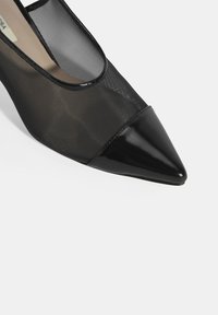Chaussure à talons hauts noire avec un bout pointu en cuir verni brillant et un dessus en maille transparente, créant un aspect texturé contrasté.