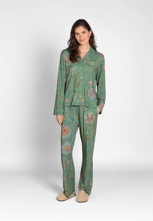 Woman wearing green floral patterned long-sleeve pajama set with relaxed fit pants and beige slippers standing against a plain background.