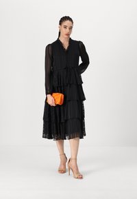 Black tiered maxi dress with long sheer sleeves, ruffled neckline, and button details, paired with an orange textured clutch and nude heels.