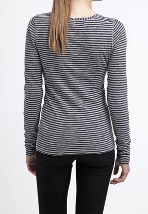 Frau mit langen Haaren, die ein eng anliegendes, langärmliges grau-schwarz gestreiftes Hemd und schwarze Hosen trägt, von hinten gezeigt.