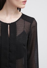 Donna che indossa una blusa trasparente nera con testo in tessuto, con piccolo bottone di chiusura al collo, che mostra la clavicola e parte del viso di profilo.