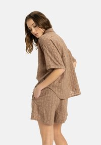Kurzärmliges, strukturiertes braunes Oberteil mit Kragen und seitlichen Taschen, kombiniert mit passender, strukturiertem Shorts. Beide besitzen ein kreisförmiges Muster.