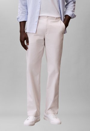 Osoba ubrana w jasnobeżowe spodnie o prostych nogawkach, białe trampki i niebiesko-białą pasiastą koszulę na białym T-shircie.