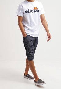 Camiseta blanca de algodón con el logo negro de "ellesse", combinada con pantalones cortos oscuros con estampado y zapatos grises slip-on con suelas blancas.