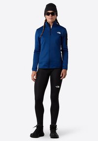 Modrá bunda na zip s dlouhými rukávy, kombinovaná s černými legínami. Venku outfit doplňují sluneční brýle a černá čepice.