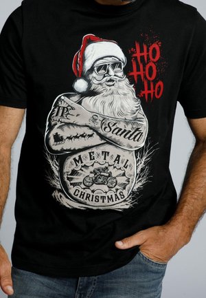 Mikołaj w okularach przeciwsłonecznych i tatuażach, z założonymi na piersi ramionami, w kapeluszu, z motocyklowym wzorem "Ho Ho Ho" i "Metal Christmas" na czarnej koszulce.