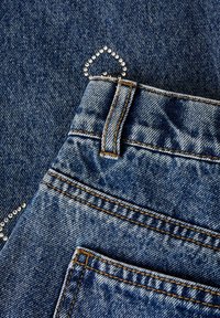 Džinsai iš mėlynos medžiagos su dekoratyviniu sidabro spalvos kniedžių raštu širdies formos priekyje šalia galinės kišenės, su oranžinėmis siūlėmis.