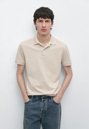 Junger Mann mit schwarzen Haaren, der ein beiges Polohemd und blaue Jeans trägt, mit den Händen in den Taschen vor einer weißen Wand stehend.