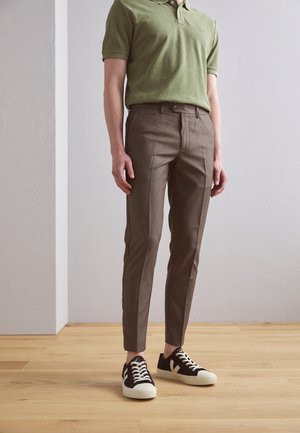 Hellgrünes Poloshirt, braune maßgeschneiderte Hose und schwarze Sneakers mit weißen Sohlen. Die Materialien wirken glatt und kombinieren formelle und lässige Stile.