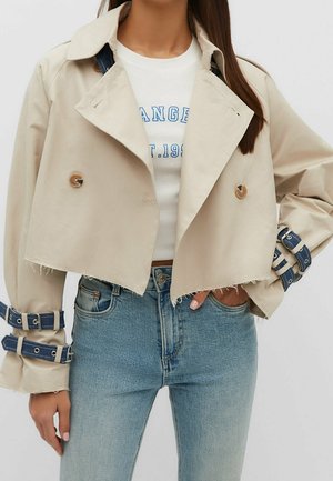 Vrouw draagt een korte beige trenchcoat met blauwe gespbanden op de mouwen, een wit bedrukt T-shirt en lichtblauwe spijkerbroek.
