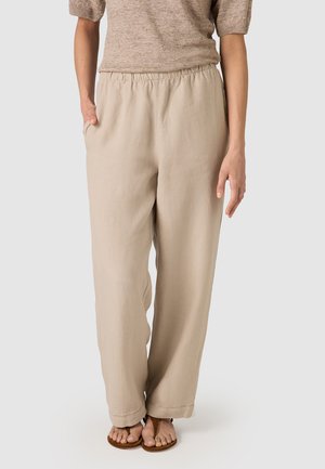 Personne portant un pantalon beige à taille élastique, jambes larges, avec poches latérales et des sandales plates marron, debout devant un fond uni.
