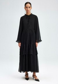 Black tiered dress with a ruffled neckline and long sleeves. Sheer fabric, fitted bodice, and textured details at the cuffs.