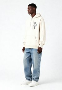 Sweat à capuche crème avec un motif de tulipe noire, poche kangourou à l'avant, capuche à cordon, associé à un jean bleu clair ample et des baskets blanches.
