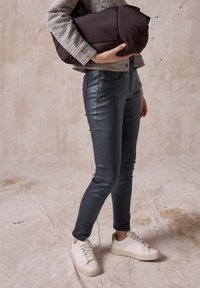 Mörka smala jeans med en slät, glänsande yta, i kombination med en mönstrad topp och vita sneakers. Håller en mörk pufferväska.