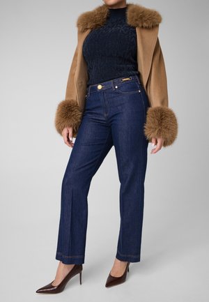 Kvinde iført mørkeblå jeans, en marineblå mønstret top, en kamelfarvet frakke med pelskrave og manchetter, og brune højhælede sko.