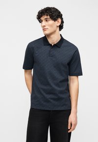 Νεαρός άνδρας με σγουρά σκούρα μαλλιά, που φοράει μπλε πόλο μπλούζα με κοντά μανίκια και μαύρο παντελόνι, στεκόμενος μπροστά σε απλό φόντο.