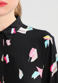 Camisa negra con cuello, que presenta un patrón de hojas colorido en rosa, azul y verde. Hecha de un tejido ligero y suave.