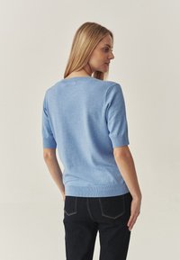 Pulover tricotat de culoare albastru deschis, cu mâneci scurte, decolteu și tiv cu dungi, textură fină, asortat cu blugi negri din denim.