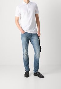 Polo blanco de manga corta con logo, combinado con jeans azul claro descoloridos y zapatos estilo mocasín negros, posando frente a un fondo neutro.