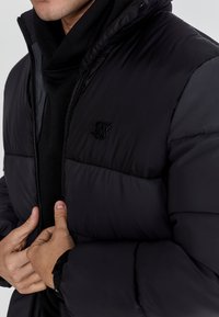 Schwarze Daunenjacke mit Reißverschluss, matte Oberfläche, hoher Kragen und einem kleinen Logo auf der Brust. Texturiertes Gewebe mit sichtbaren Nähten.