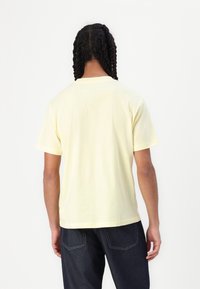 Osoba z warkoczami, ubrana w jasnożółtą koszulę z krótkim rękawem i ciemnoniebieskie dżinsy, stojąca plecami do kamery na jednolitym tle.