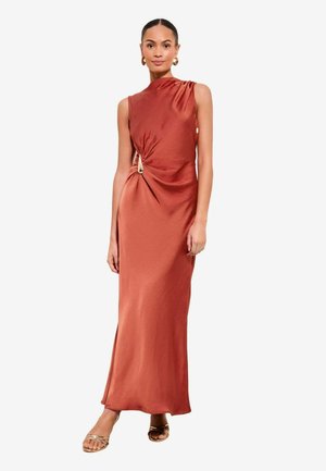 Kobieta w bezrękawną suknię z satyny w kolorze rdzy z marszczeniem z boku i złotym pierścieniem, do tego złote sandały na otwartym palcu i kolczyki-koła.