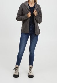 Kobieta w szarej polarowej kurtce na zamek, ciemnoniebieskich dżinsach, beżowych botkach sznurowanych i czarnej bluzce, stojąca na tle jasnego, jednolitego tła.