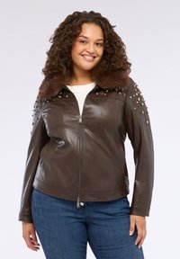 Giacca di pelle marrone con colletto in pelliccia sintetica, spalle borchiate e chiusura con zip. Presenta una texture liscia e un design aderente.