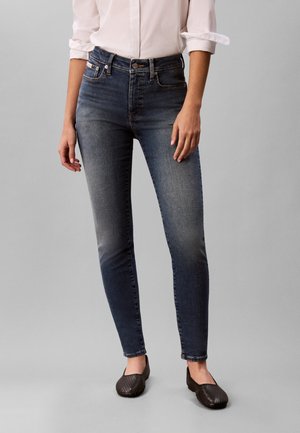 Personne portant un jean skinny bleu foncé, une chemise blanche à manches longues retroussées et des chaussures noires sans lacets debout sur un sol gris.