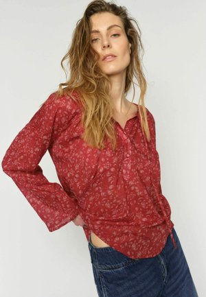 Femeie cu părul lung și ondulat, purtând o bluză roșie cu nasturi și model și blugi albaștri, pozând cu o mână pe șold pe un fundal simplu.