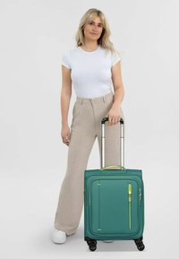 Valise roulante teal avec une surface texturée, des zips jaunes et plusieurs compartiments. Roues et poignée rétractable pour un transport facile.