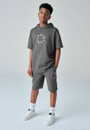 Dreng stående med hænder delvist i lommerne, iført grå hættetrøje og shorts sæt med pink cirkulær tekst, hvide sokker og hvide sneakers.