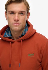 Rust-colored hoodie with adjustable drawstrings, featuring a green logo on the front and a blue plaid shirt collar layered underneath.