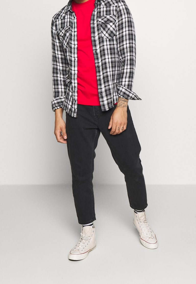 Homme portant un t-shirt rouge, une chemise à carreaux noire et blanche, un pantalon noir, des chaussettes rayées et des baskets montantes blanches, debout devant un fond uni.