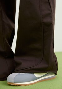 Černé sportovní kalhoty částečně zakrývají modrošedý sneaker s bílým swoosh a světlým hnědým podešví, který má texturovaný svršek a hladký povrch.