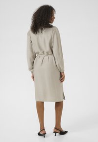 Robe beige clair à manches longues et poignets élastiques, taille ceinturée et coupe droite. Tissu texturé avec fente sur le côté. Sandales à talons noires.