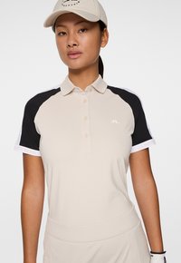 Beige golfpiké med svarta och vita detaljer, knäppning framtill och logotyp på bröstet. Slät tygkvalitet, figursydd design och korta ärmar.